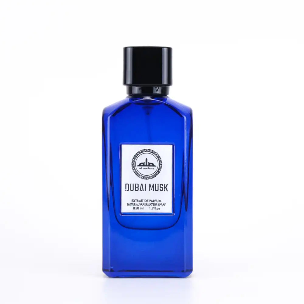 A rectangular cobalt blue glass perfume bottle by Al Ambra Perfumes, topped with a thick black cap. Its central white label features the Al Ambra Perfumes logo and "DUBAI MUSK" in prominent gold text, with "Extrait de Parfum 50 ml / 1.7Fl.oz." detailed below. The bottle stands on a reflective white background.